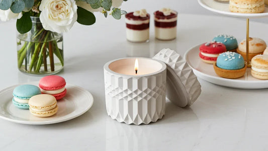 Handgemachte Kerze in weißem, strukturiertem Glas auf Tisch mit Macarons und Blumen, stimmungsvolles Licht