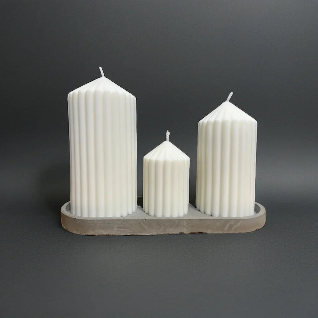 Drei handgemachte weiße Stumpenkerzen mit Rillenstruktur auf grauem Tablett, dekorativ arrangiert.