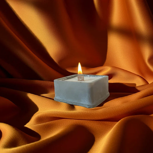 Brennende handgemachte Kerze im eckigen Behälter auf orangefarbenem Stoff, stimmungsvolles Licht