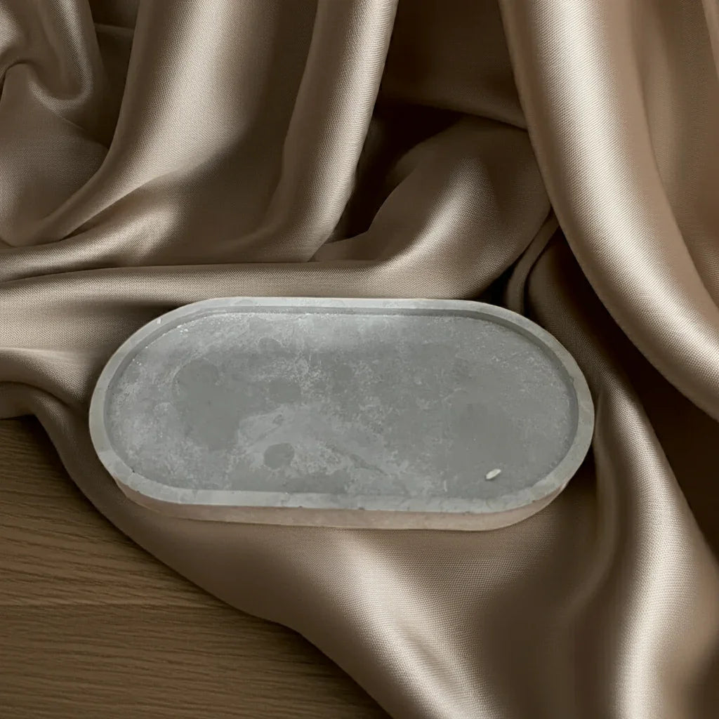 Graues, ovales Kerzentablett aus Stein auf beigem, glänzendem Stoff drapiert.