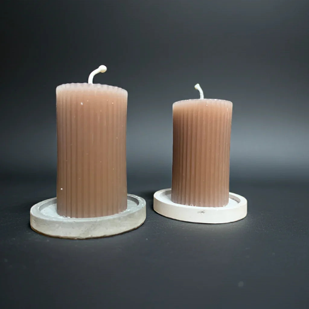 Zwei handgemachte braune Stumpenkerzen mit Rillenstruktur auf weißen Untersetzern vor schwarzem Hintergrund.