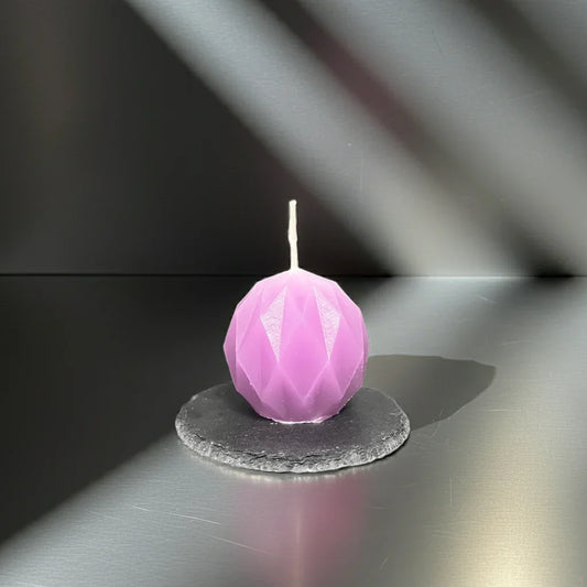 Handgemachte, lila Kugelkerze mit Facetten-Design auf Schieferplatte im Sonnenlicht